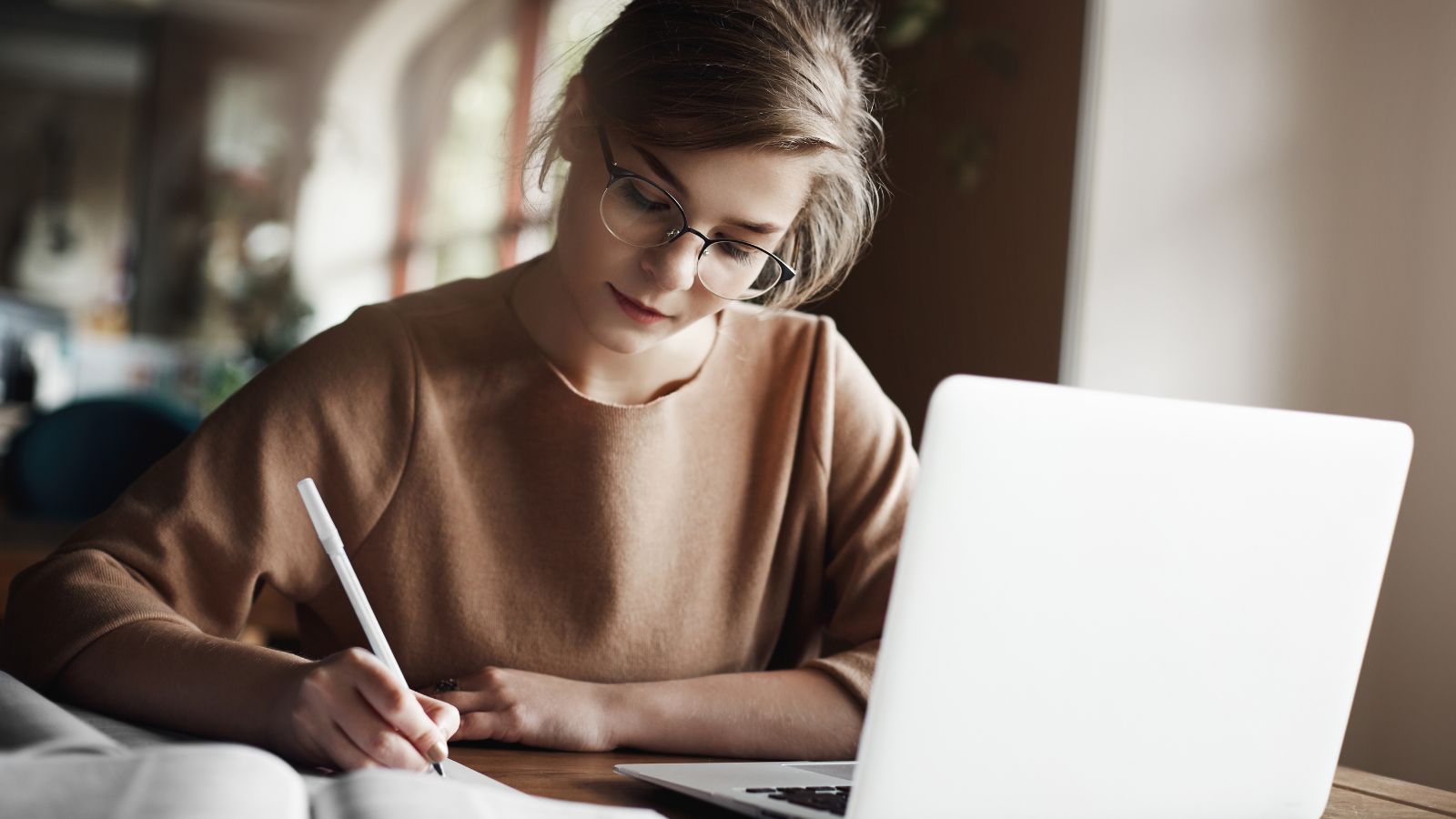 Mulher branca e loira escrevendo em um papel e seu notebook ao lado