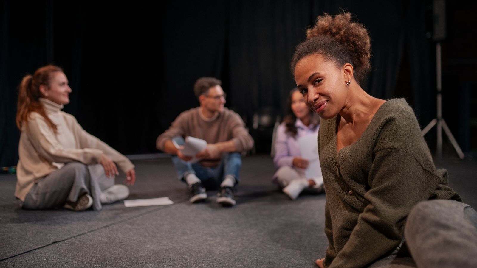 Mulher negra em destaque sentada num palco, com três pessoas desfocadas ao fundo.