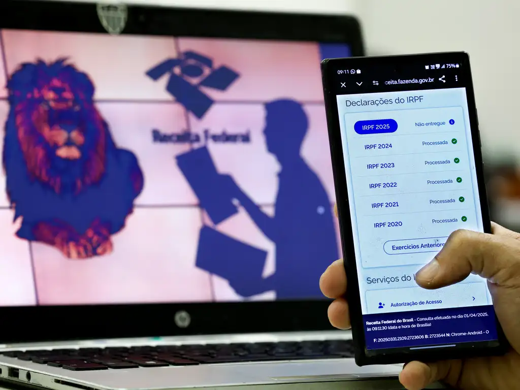 Mão segurando um celular com a tela no site da Receita Federal para declaração de Imposto de Renda 2025, com computador ao fundo.