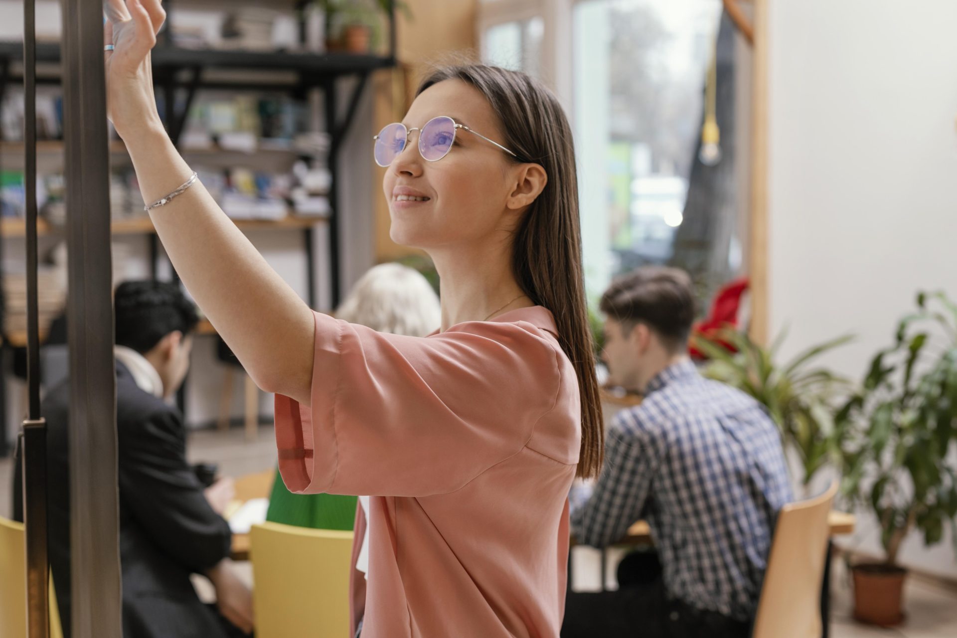 Mulher em escritório anotando em quadro, com colegas de trabalho ao fundo